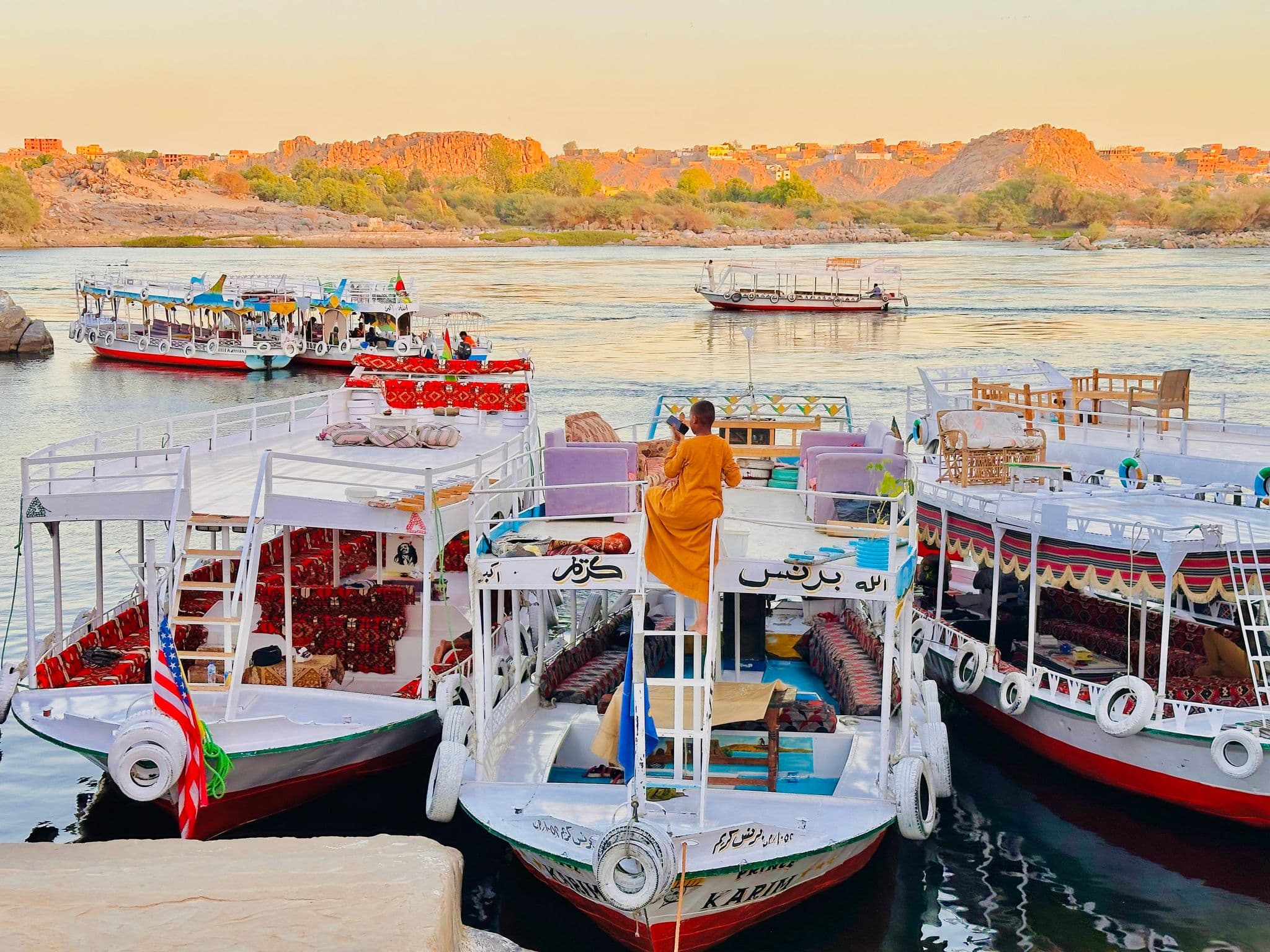 Colorful riverboats moored on the Nile River in Aswan, Egypt, with a person in an orange robe climbing a ladder on one boat.