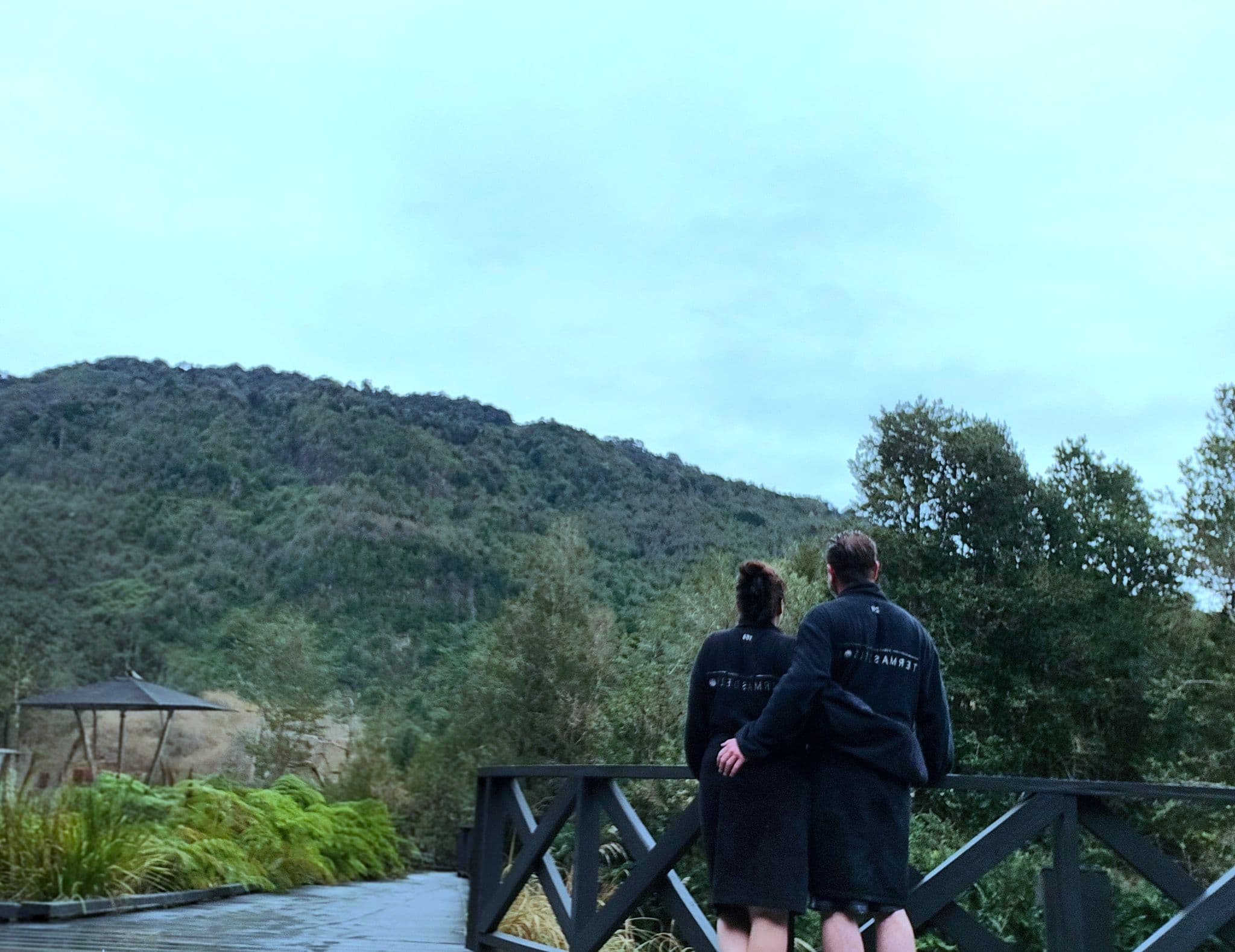 Two people in black spa robes standing on a wooden bridge, looking toward forested hills near hot springs in Chile.