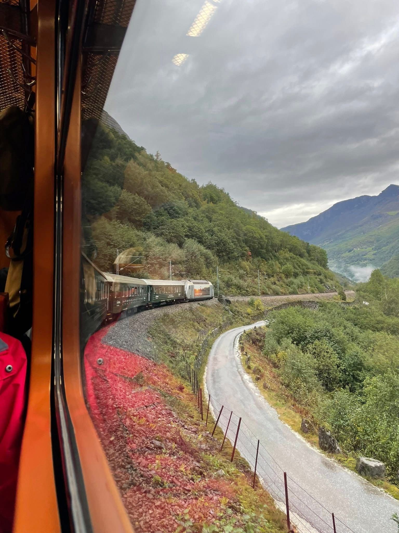 Flåm Railway train winding along a mountainside, seen through a carriage window with a valley road and green hills, Norway.