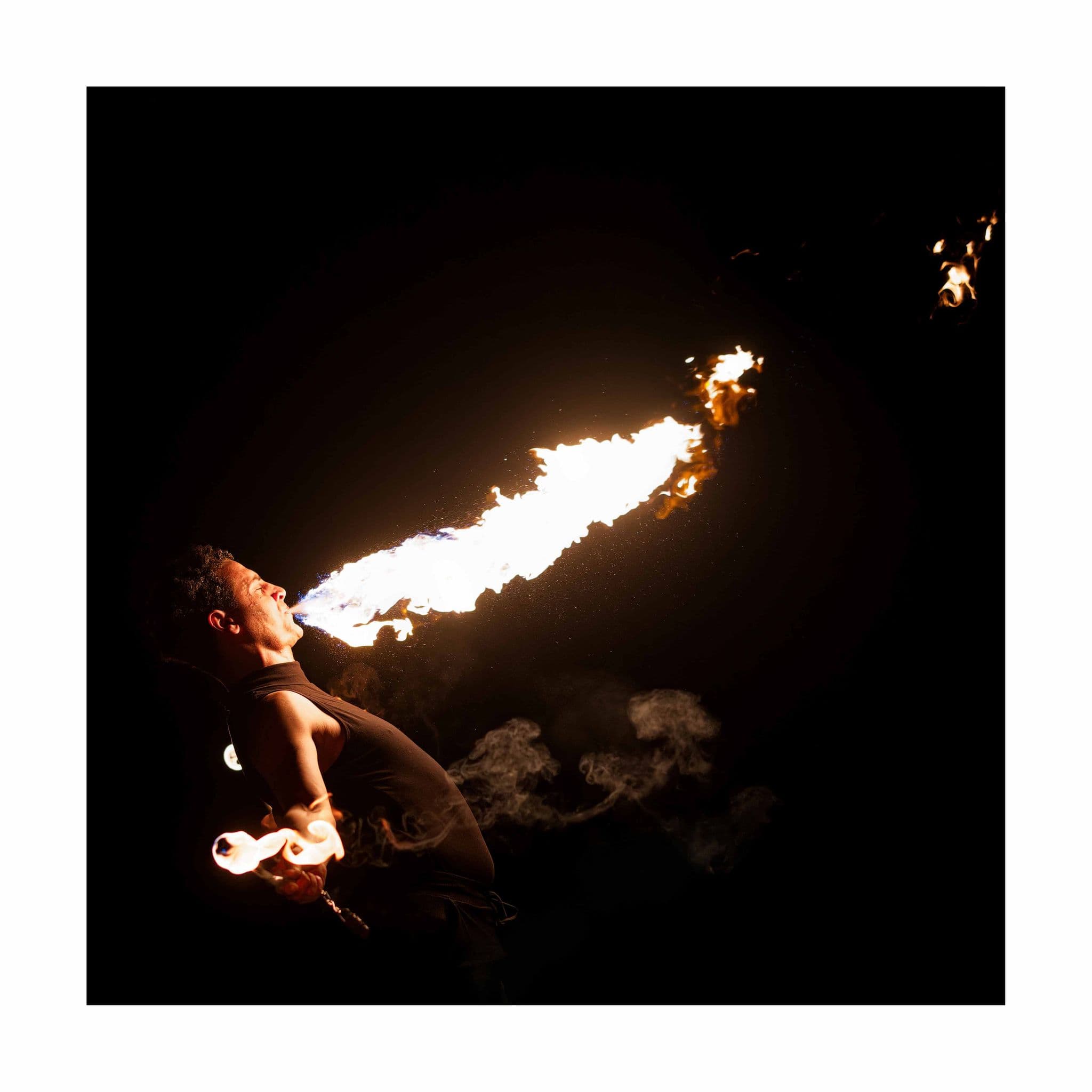 A fire eater blowing a long plume of flame during a nighttime outdoor performance, smoke trailing into the dark.