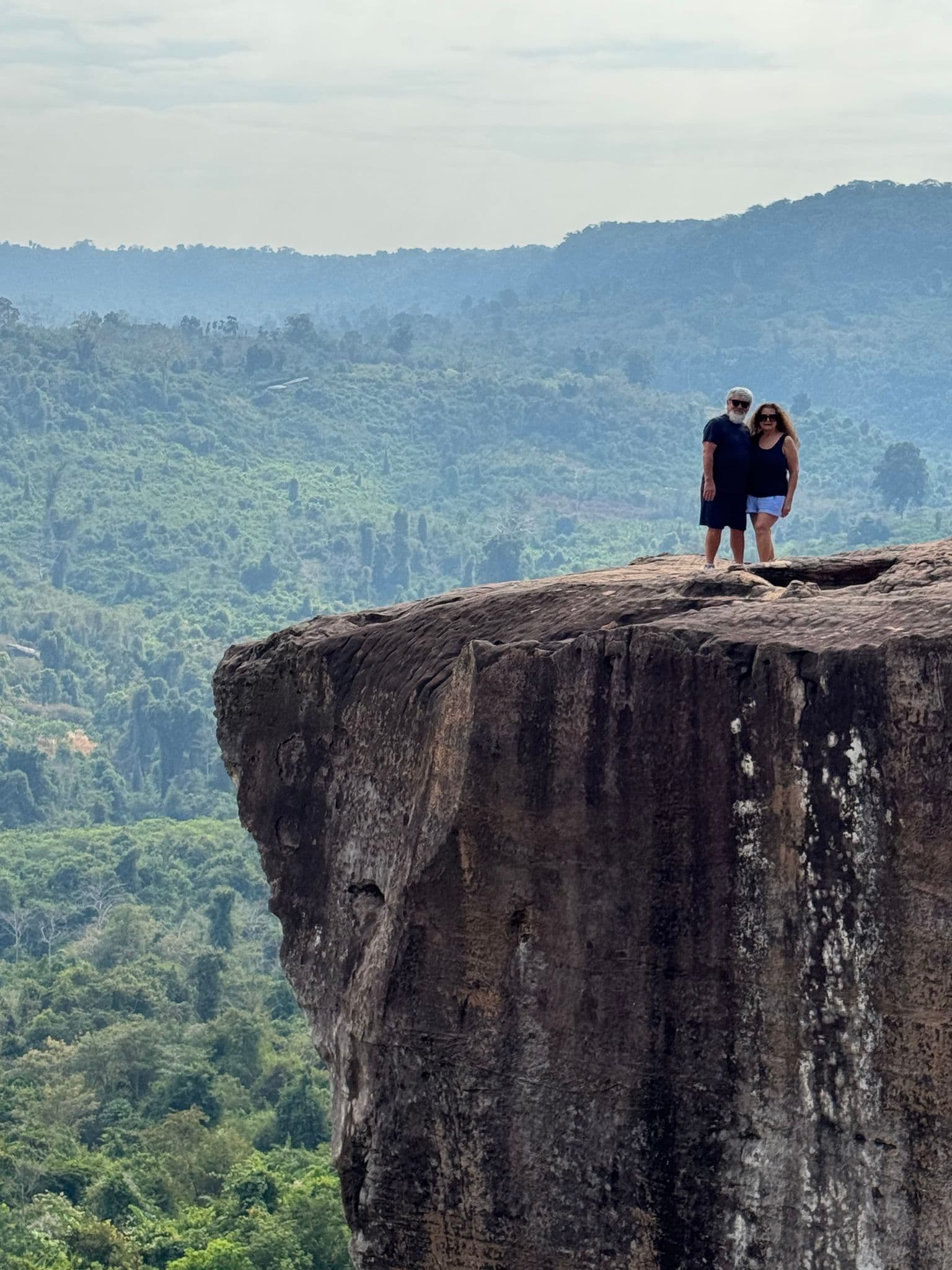 Cliff at Phnom Kulen with two travelers standing at the rocky edge overlooking forested hills in Cambodia.