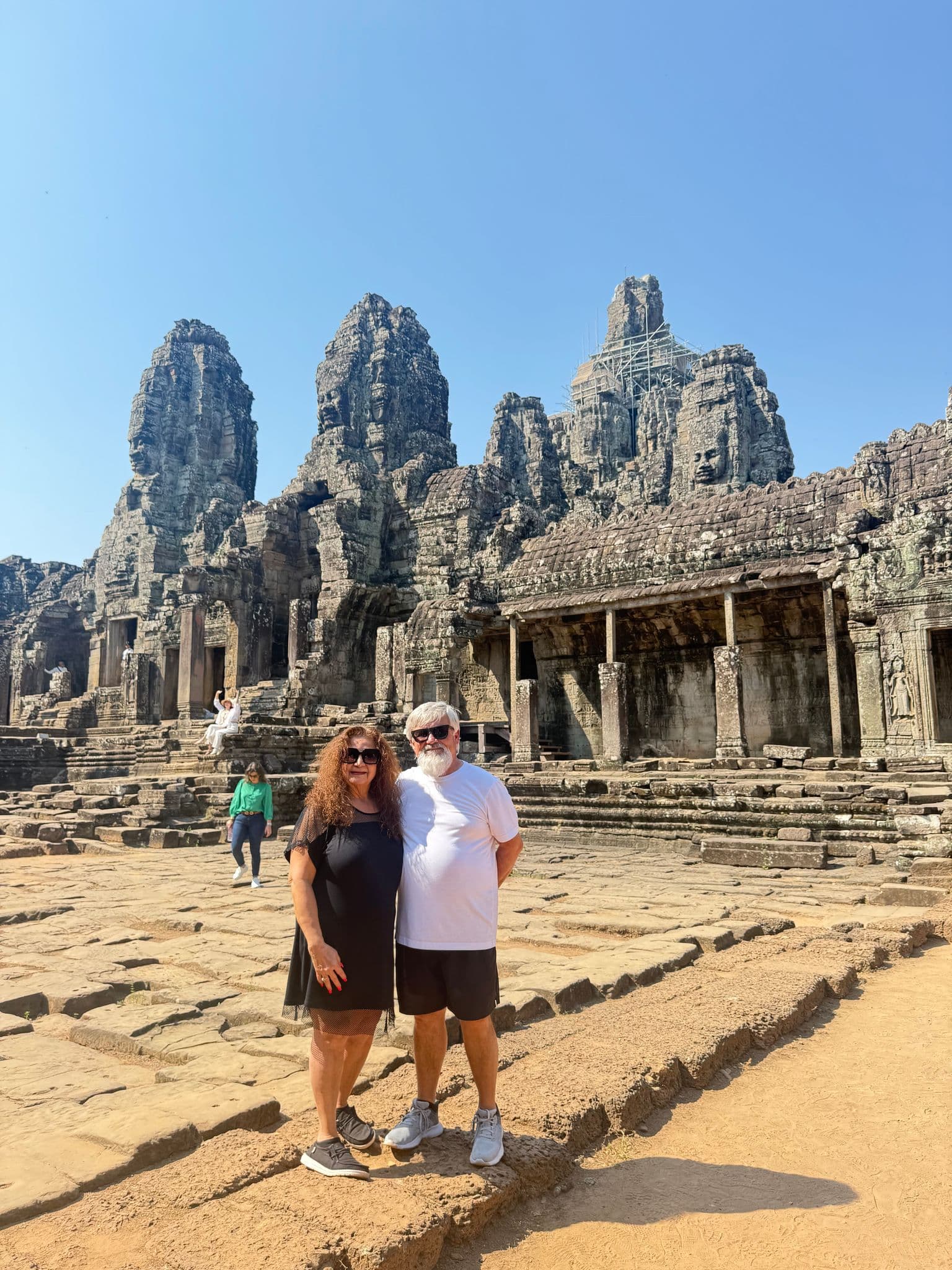 Stone face towers of Bayon Temple in Angkor Thom, Cambodia, with two tourists standing on the temple courtyard.