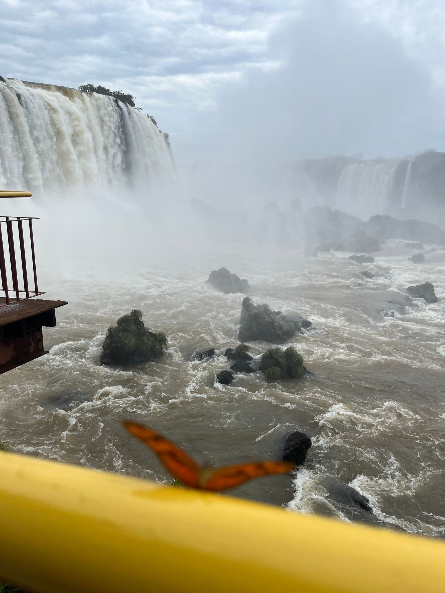 Iguazu Falls cascading over cliffs with heavy mist and rocky rapids, a blurred orange butterfly perched on a yellow railing in the foreground.