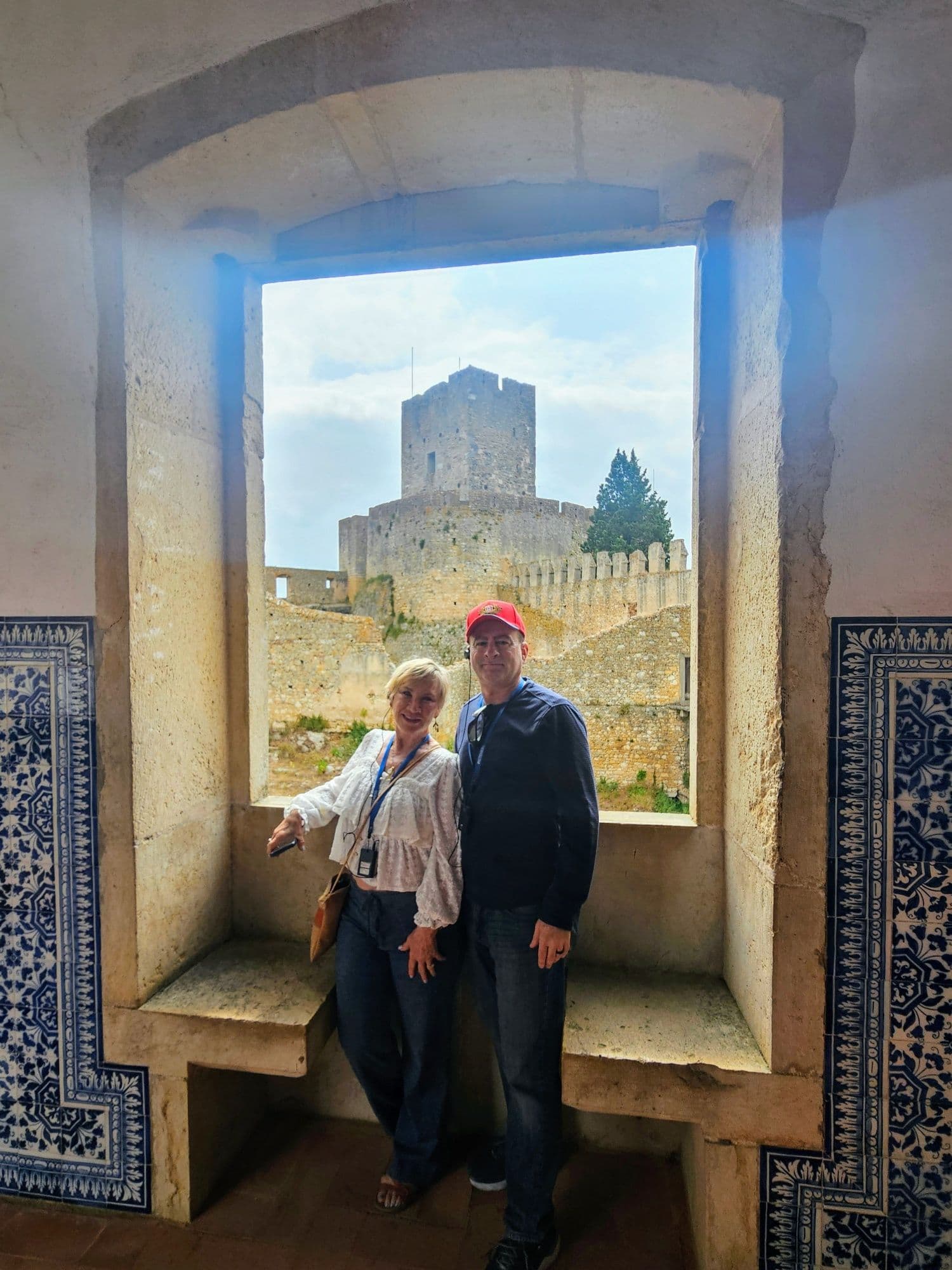 Stone castle tower seen through a large window with a couple standing on the inner sill inside a historic castle in Portugal.