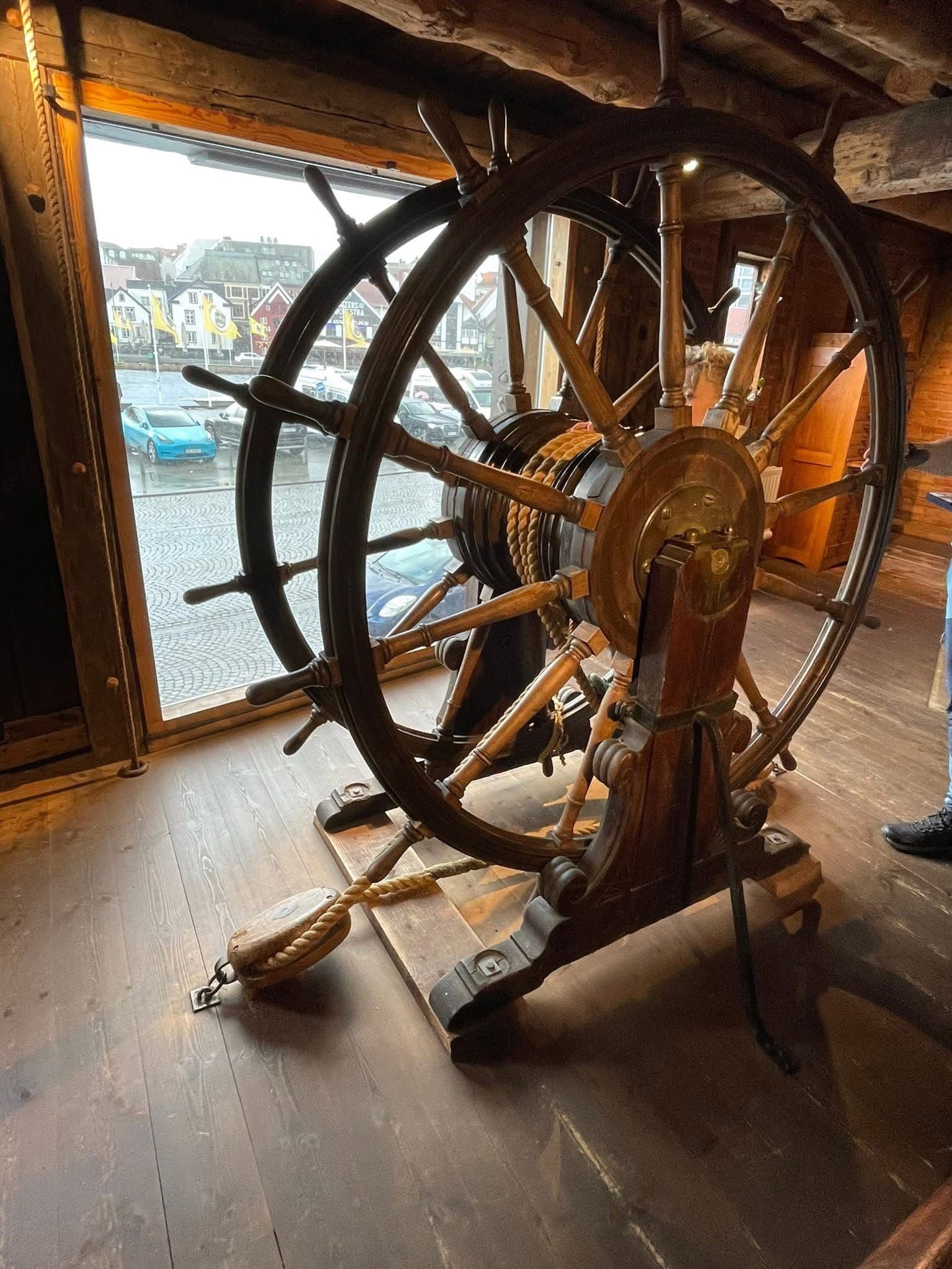 Wooden ship's wheel and capstan inside Stavanger Maritime Museum, with the harbor and waterfront buildings visible outside, Stavanger, Norway.