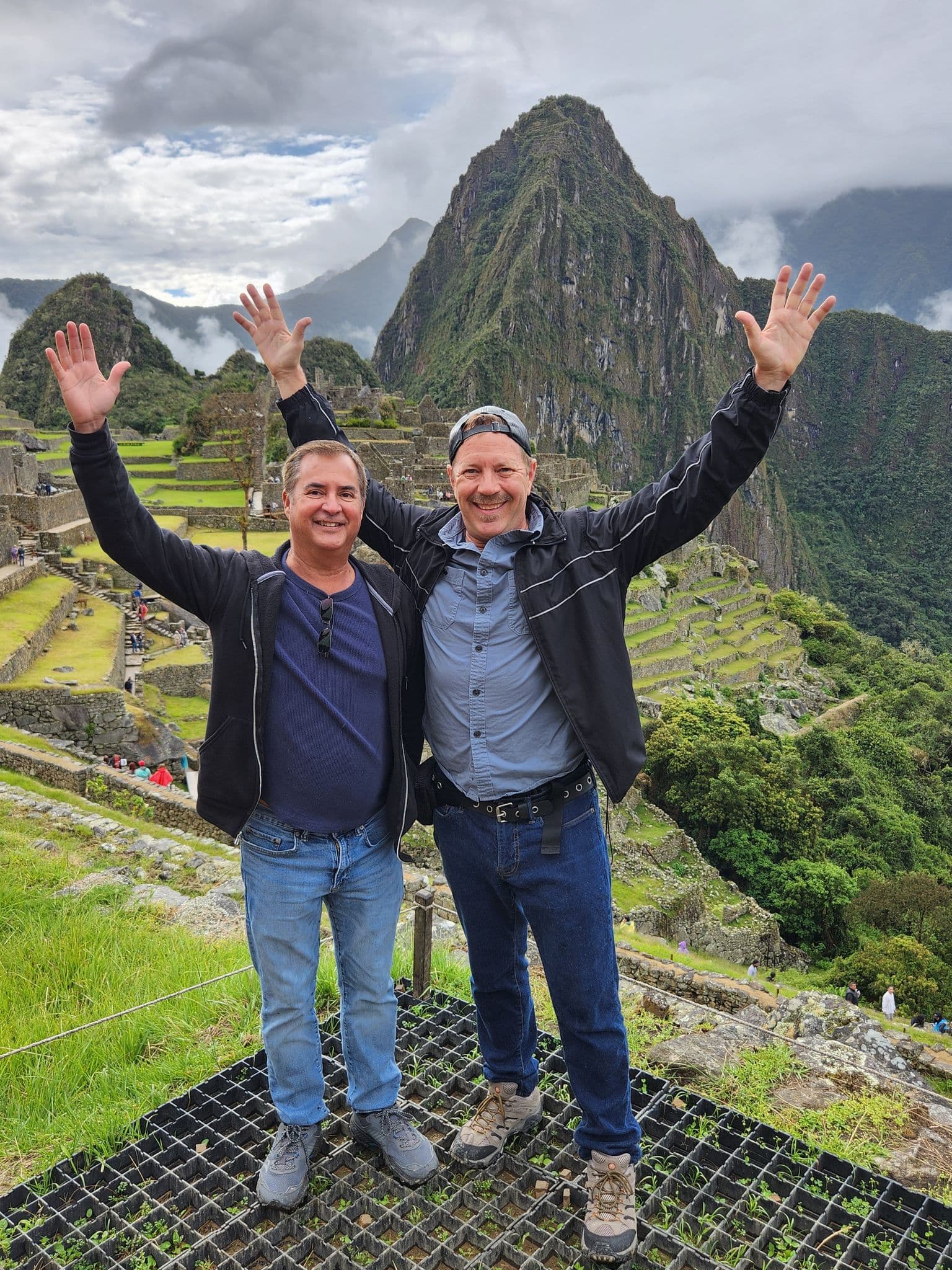 Machu Picchu terraces with two travelers standing in the foreground raising their arms, Cusco region, Peru.