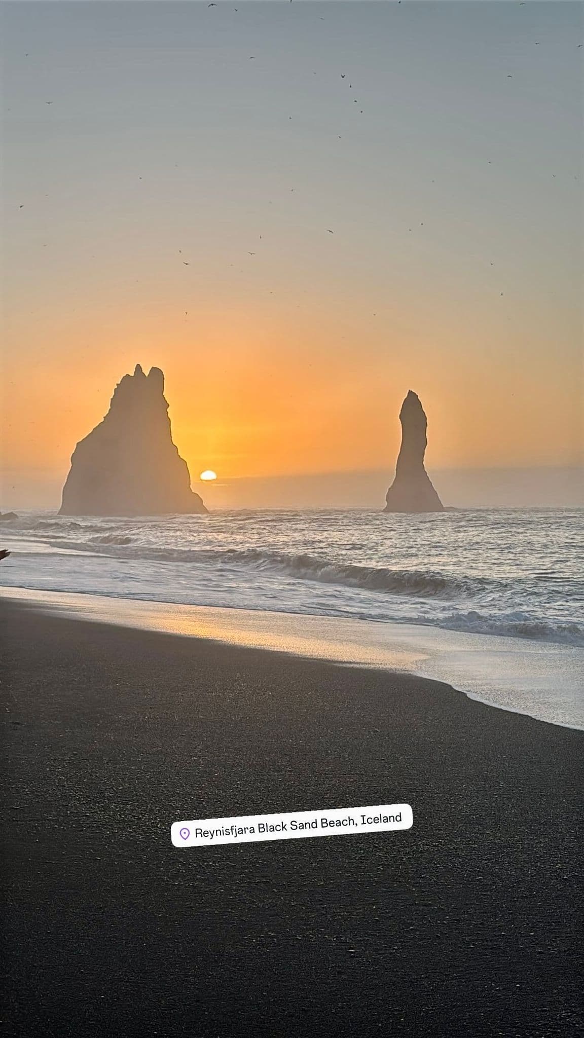 Reynisdrangar sea stacks rising from the ocean with the sun setting over Reynisfjara Black Sand Beach, Iceland.