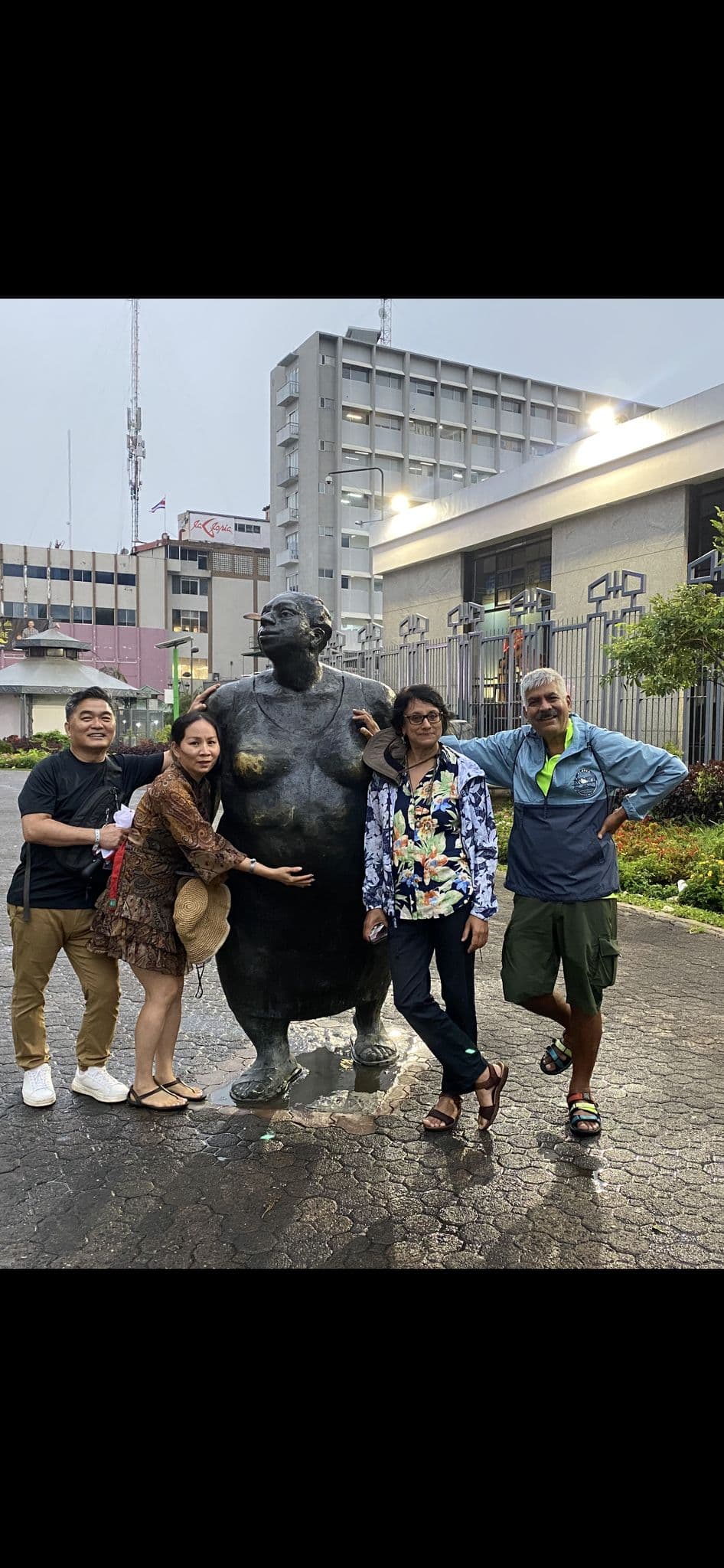 Bronze rotund sculpture with four travelers posing around it on a wet city plaza, buildings and a Thai flag visible, Thailand.