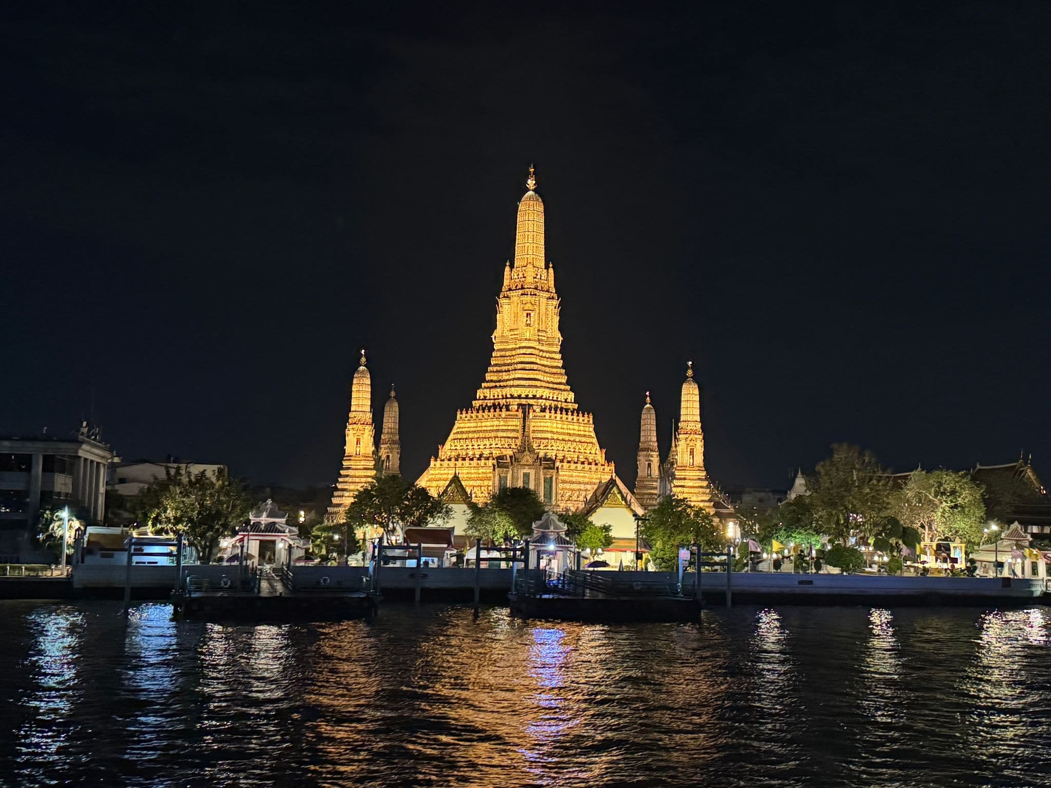Wat Arun temple lit up at night on the Chao Phraya River in Bangkok, Thailand, with lights reflecting on the water.