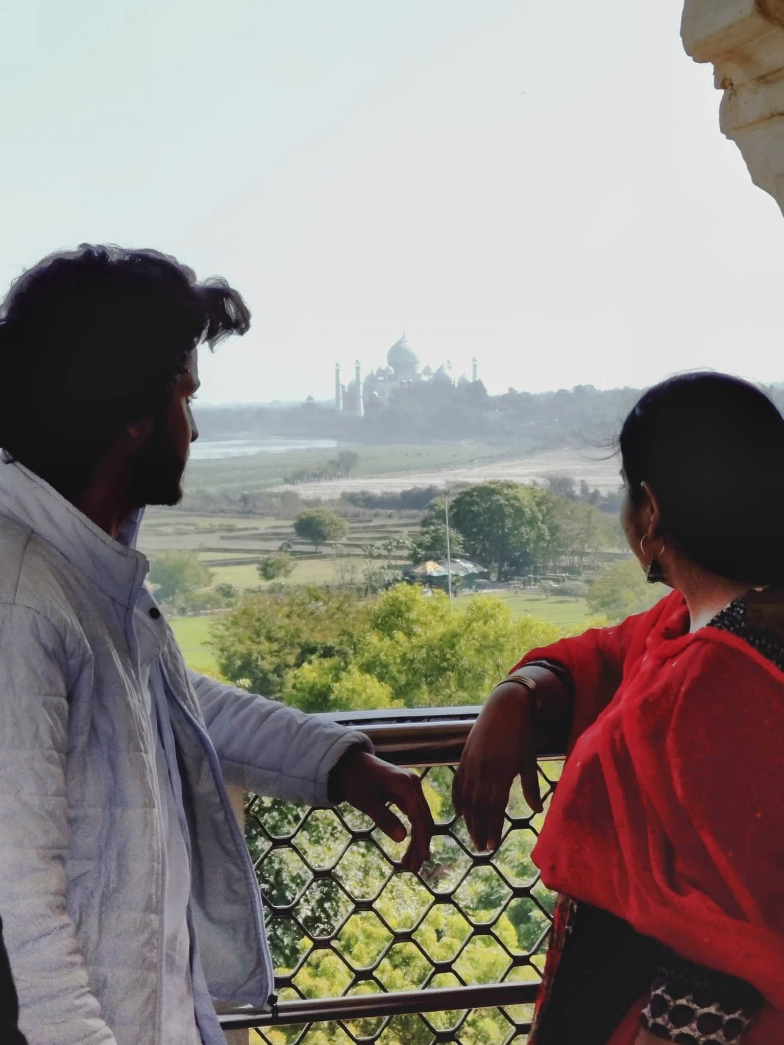 Taj Mahal framed across the river as two people lean on a balcony railing and look toward it, Agra, India.