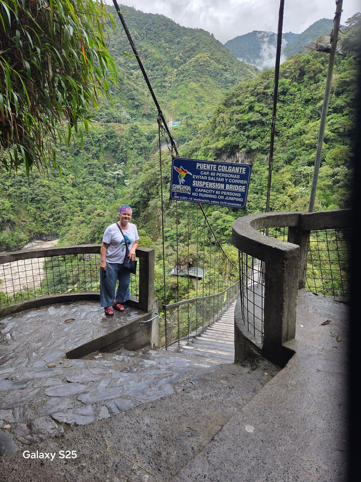 Suspension bridge entrance with a traveler standing on a stone lookout above a green Andean valley, Puente Colgante, Peru.