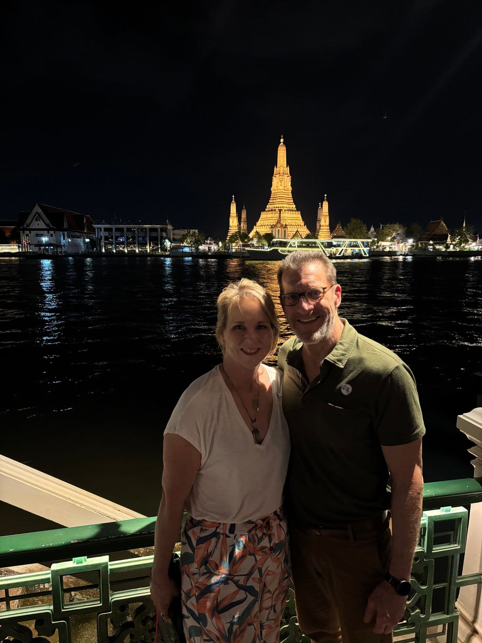 Wat Arun temple lit at night across the Chao Phraya River, with a couple posing on a riverside railing in Bangkok, Thailand.