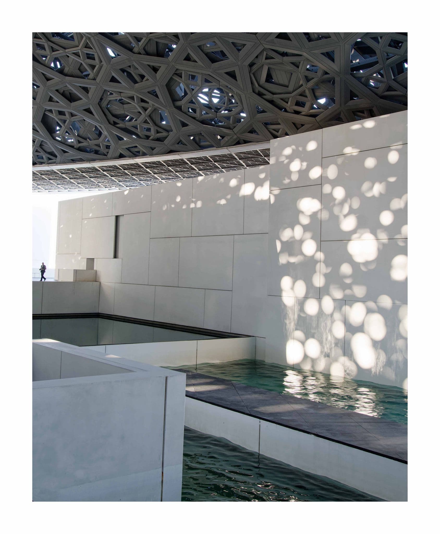 Louvre Abu Dhabi dome casting dappled light on white gallery walls and reflecting pools, with a person on a walkway, Abu Dhabi, United Arab Emirates.