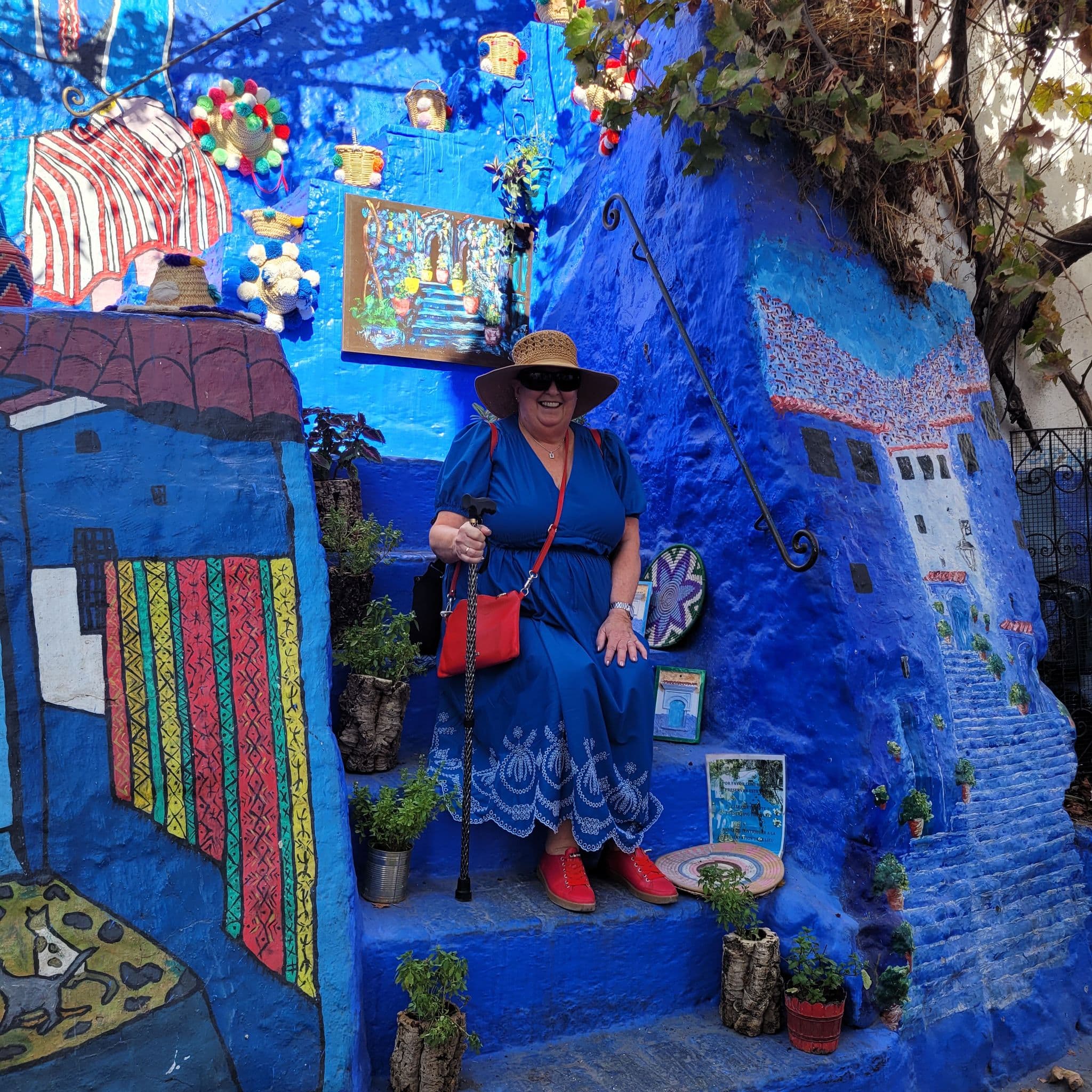 Blue-painted steps and murals in Chefchaouen, Morocco, with a woman in a blue dress and sunhat seated on the steps.