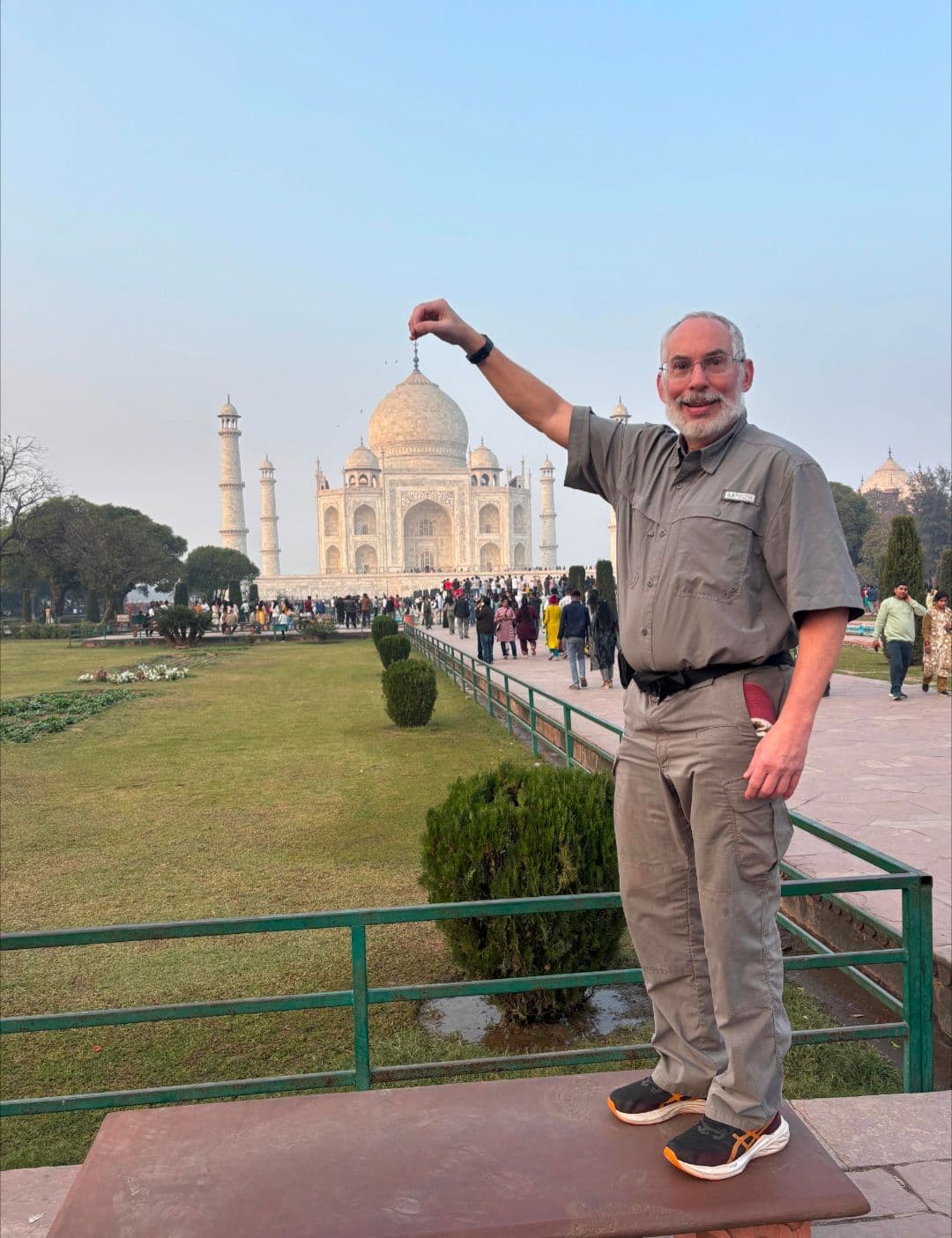 Taj Mahal in Agra, India with a man posing to pinch the dome while crowds walk the gardens.