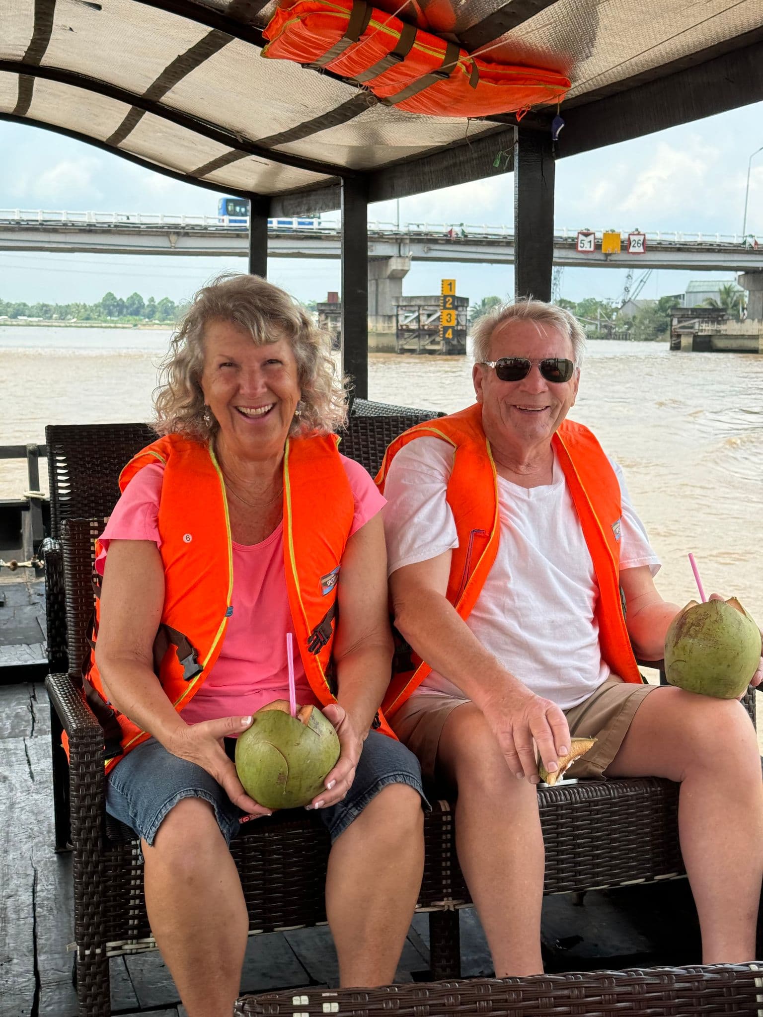 Mekong River boat with two travelers wearing orange life jackets holding coconuts on a covered cruise in the Mekong Delta, Vietnam.