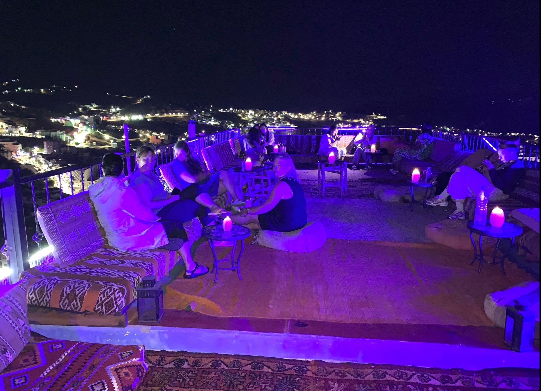 Rooftop terrace with travelers sitting around low tables and glowing lanterns, overlooking Chefchaouen at night, Morocco.