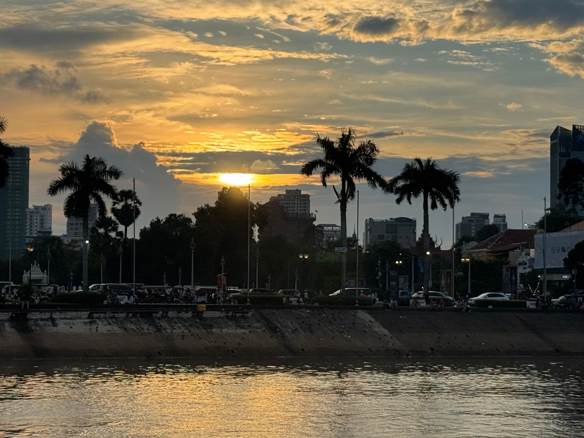 Sunset over the river and palm-lined Sisowath Quay in Phnom Penh, Cambodia, with cars and people along the promenade.