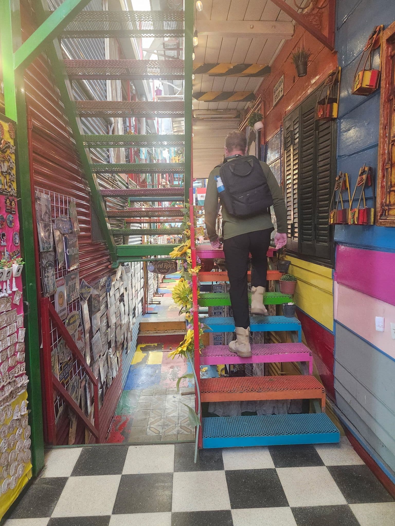 A person walking up rainbow-colored metal stairs inside a narrow market corridor lined with stalls.