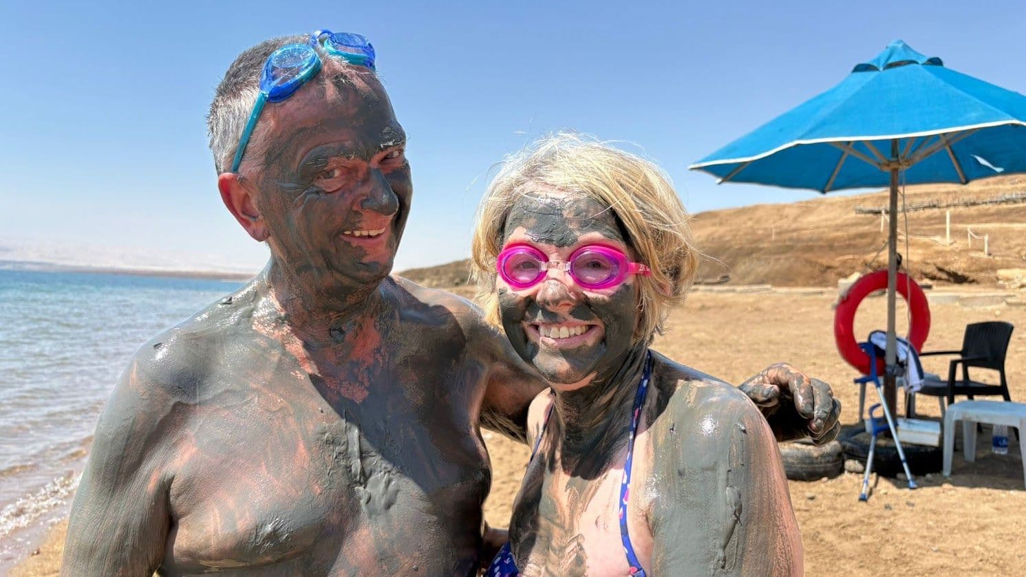 Two people covered in Dead Sea mud smiling on the shoreline of the Dead Sea after a buoyant swim.