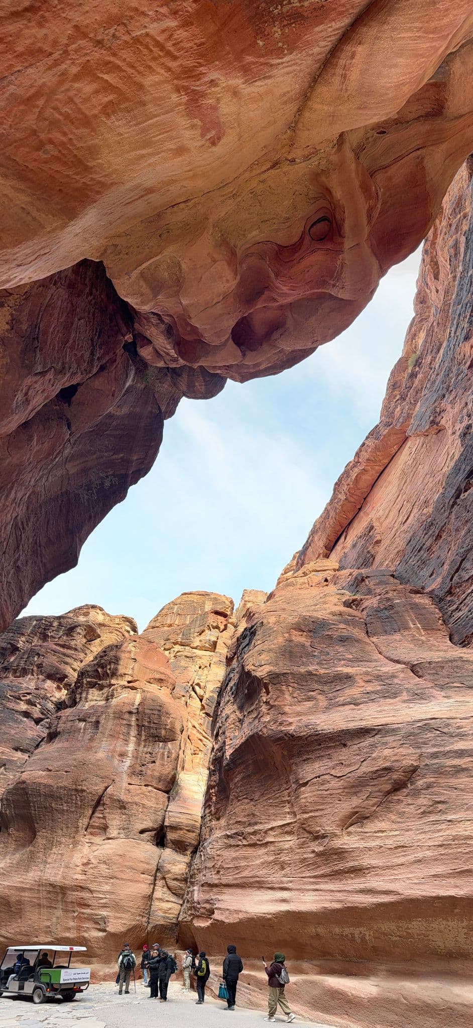 Tall red sandstone canyon walls of the Siq at Petra, Jordan, with visitors and a small tour vehicle on the canyon floor.