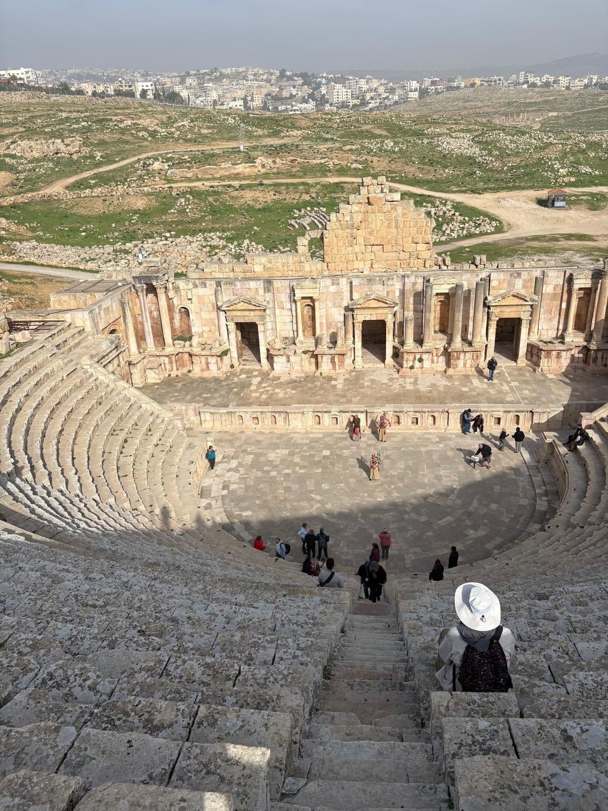 Roman theater seating and stage at Jerash (Gerasa), Jordan, seen from high steps with visitors and a person in a white hat.