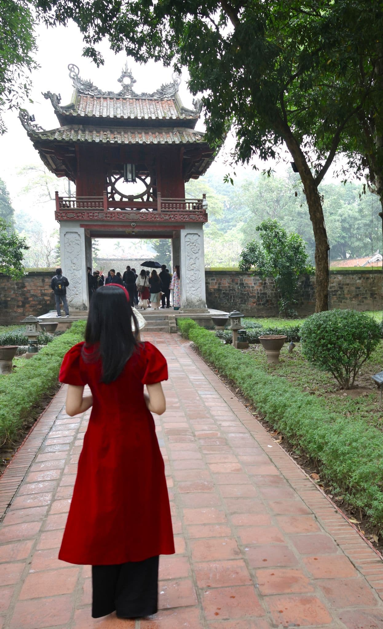Temple of Literature gate in Hanoi with a person in a red dress walking toward the entrance where a group is gathered, Vietnam.