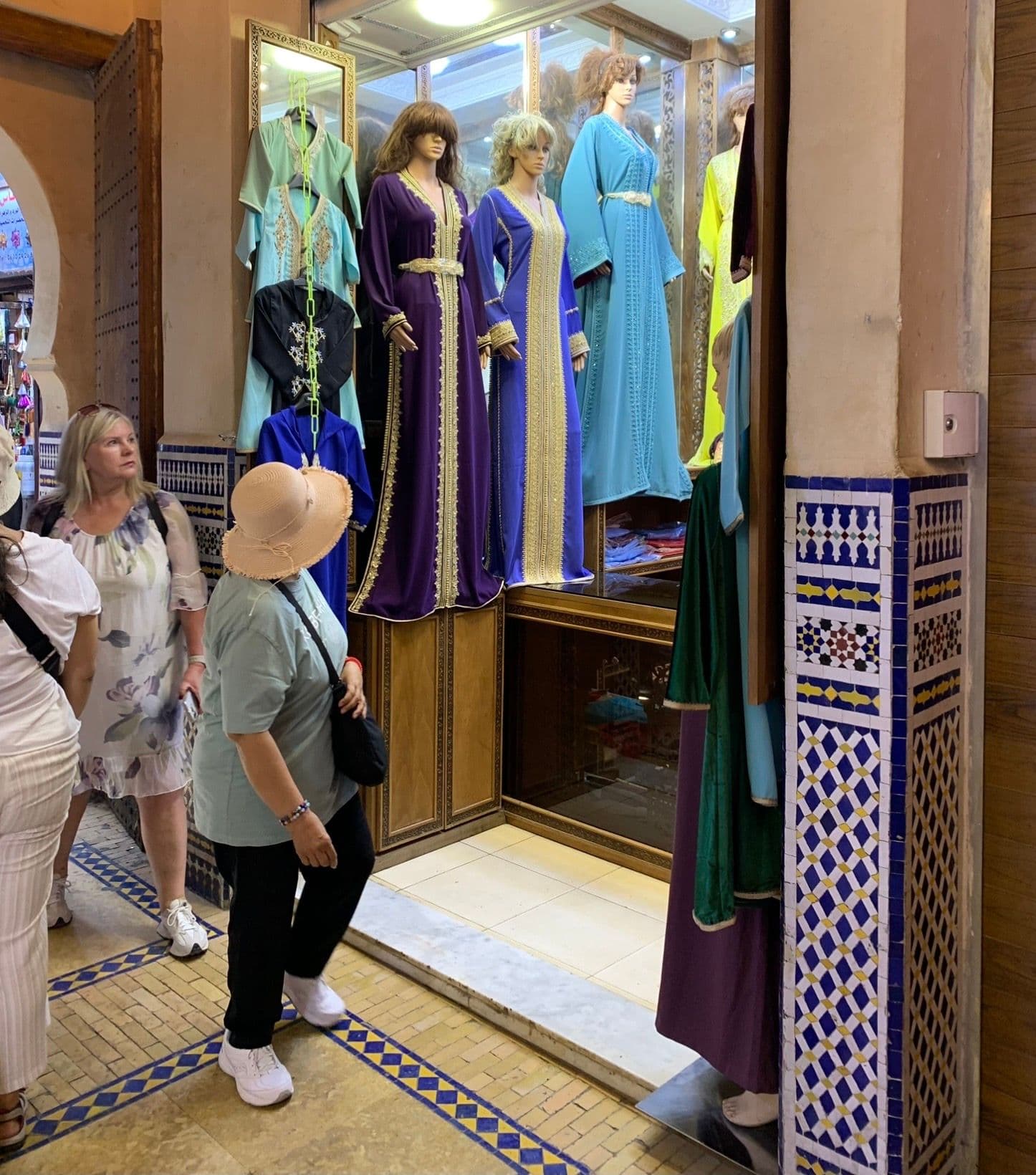 Colorful caftans on mannequins in a shop window in the Marrakech souk, with tourists browsing beside decorative mosaic tiles, Morocco.