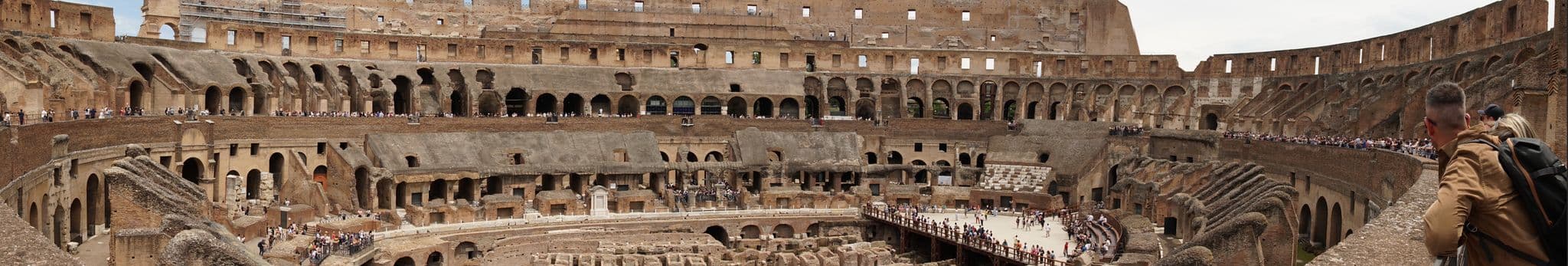Colosseum interior, Rome, Italy, with tourists on the arena floor and a visitor leaning on an upper railing.