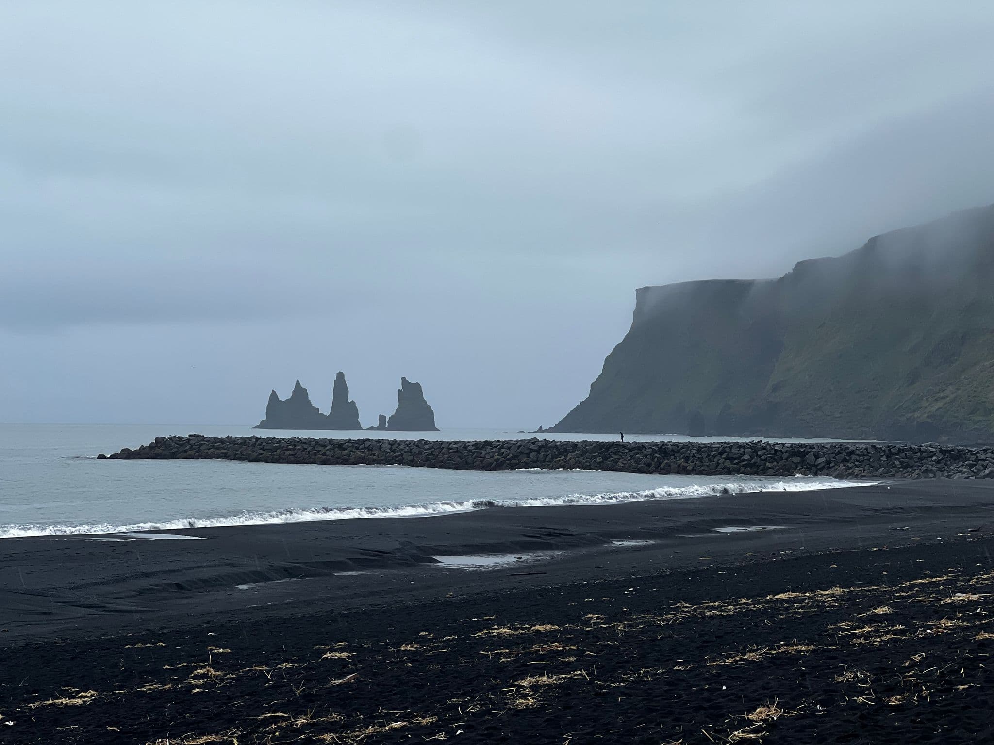 Reynisdrangar sea stacks off the black sand beach at Vík, Iceland, with waves and a lone person on a rock jetty.