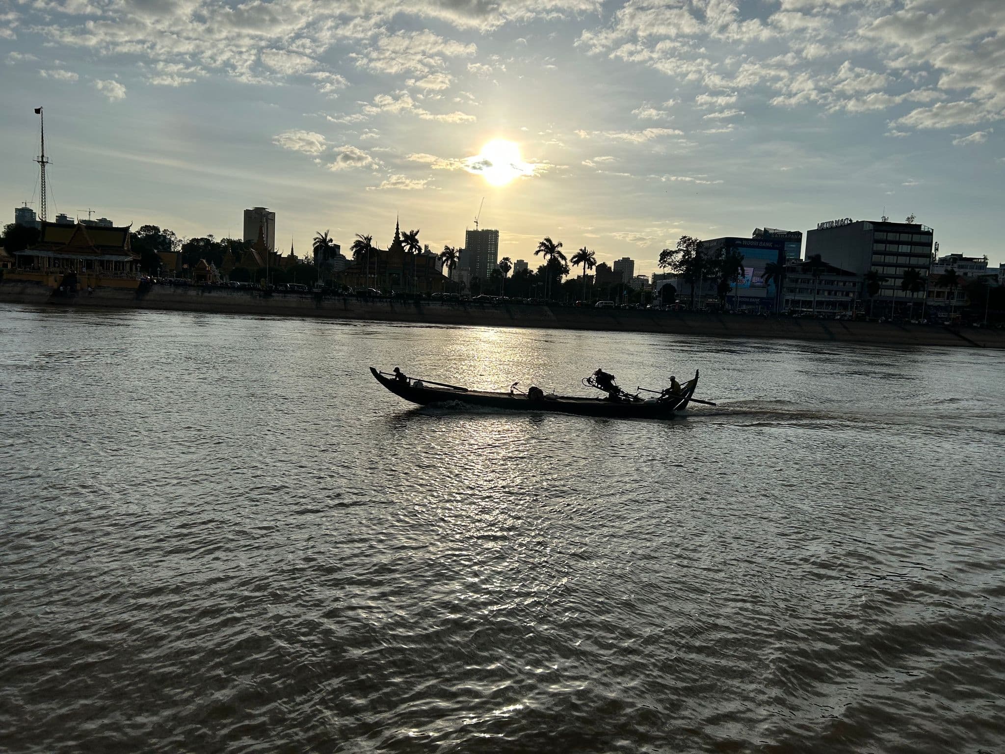 Long wooden boat silhouetted on the Tonle Sap River with Phnom Penh riverside (Sisowath Quay) and a setting sun.