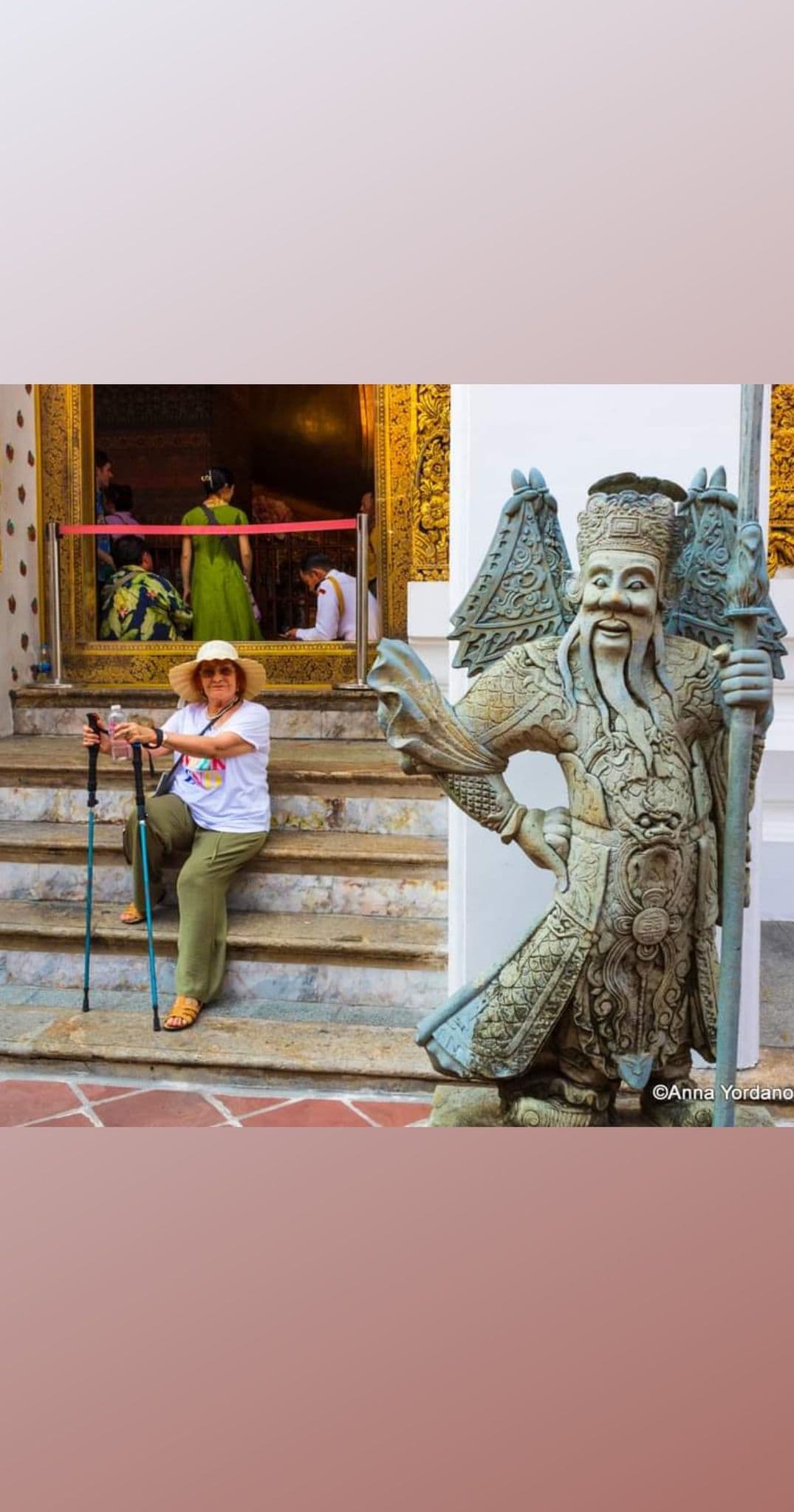 Stone guardian statue at Wat Phra Kaew, Bangkok, Thailand, beside a seated tourist with walking poles on temple steps.