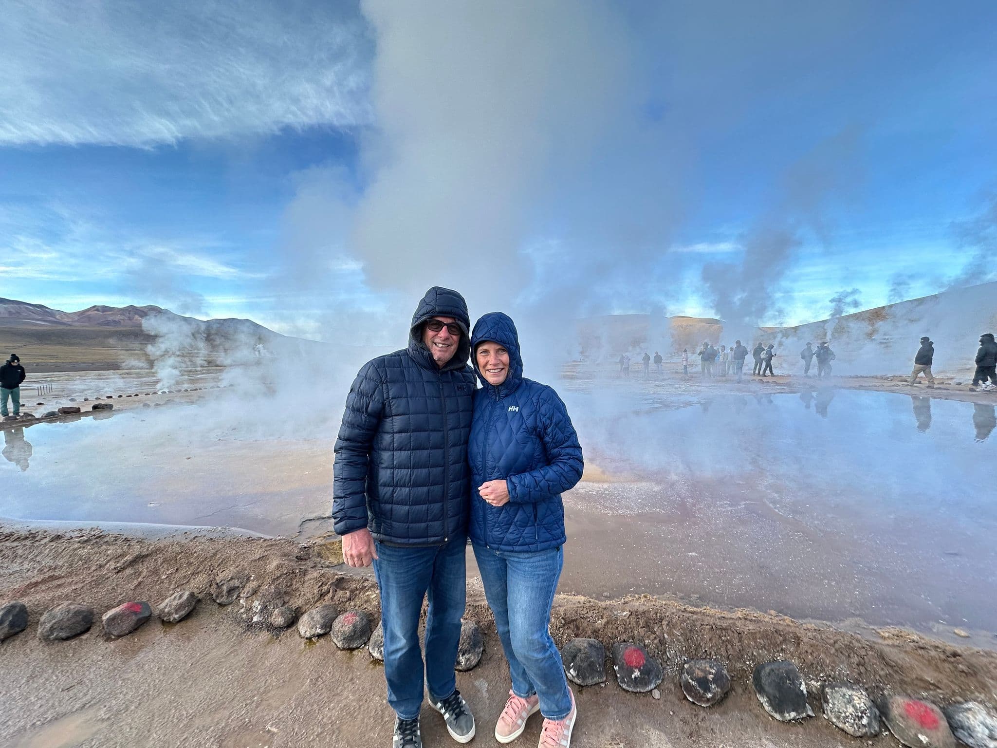 El Tatio geysers with tall steam columns and a couple posing at the edge of a steaming pool in the Chilean Altiplano, Chile.