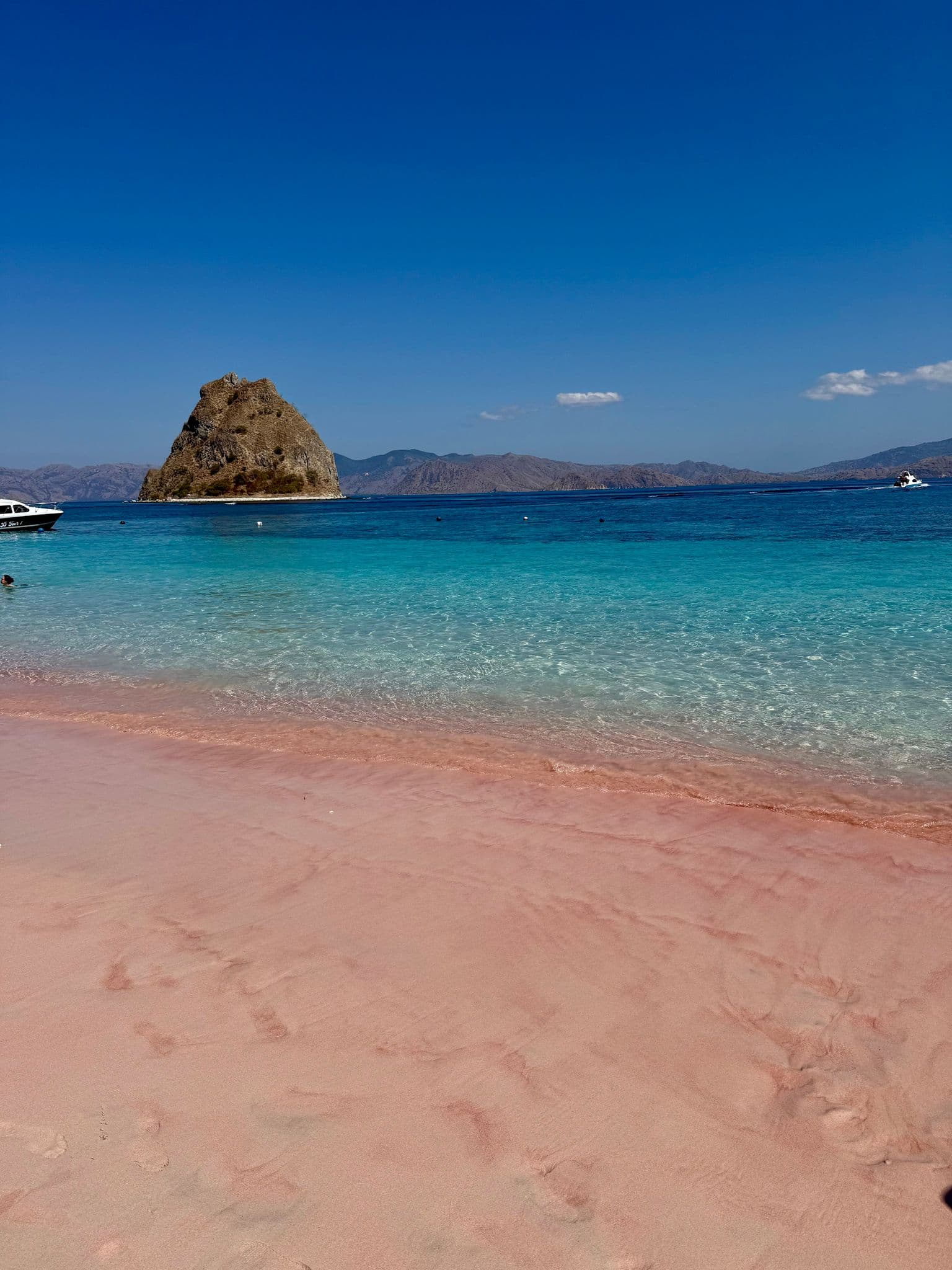 Pink sand shoreline with clear turquoise water and a rocky islet at Pink Beach (Pantai Merah), Komodo National Park, Indonesia.