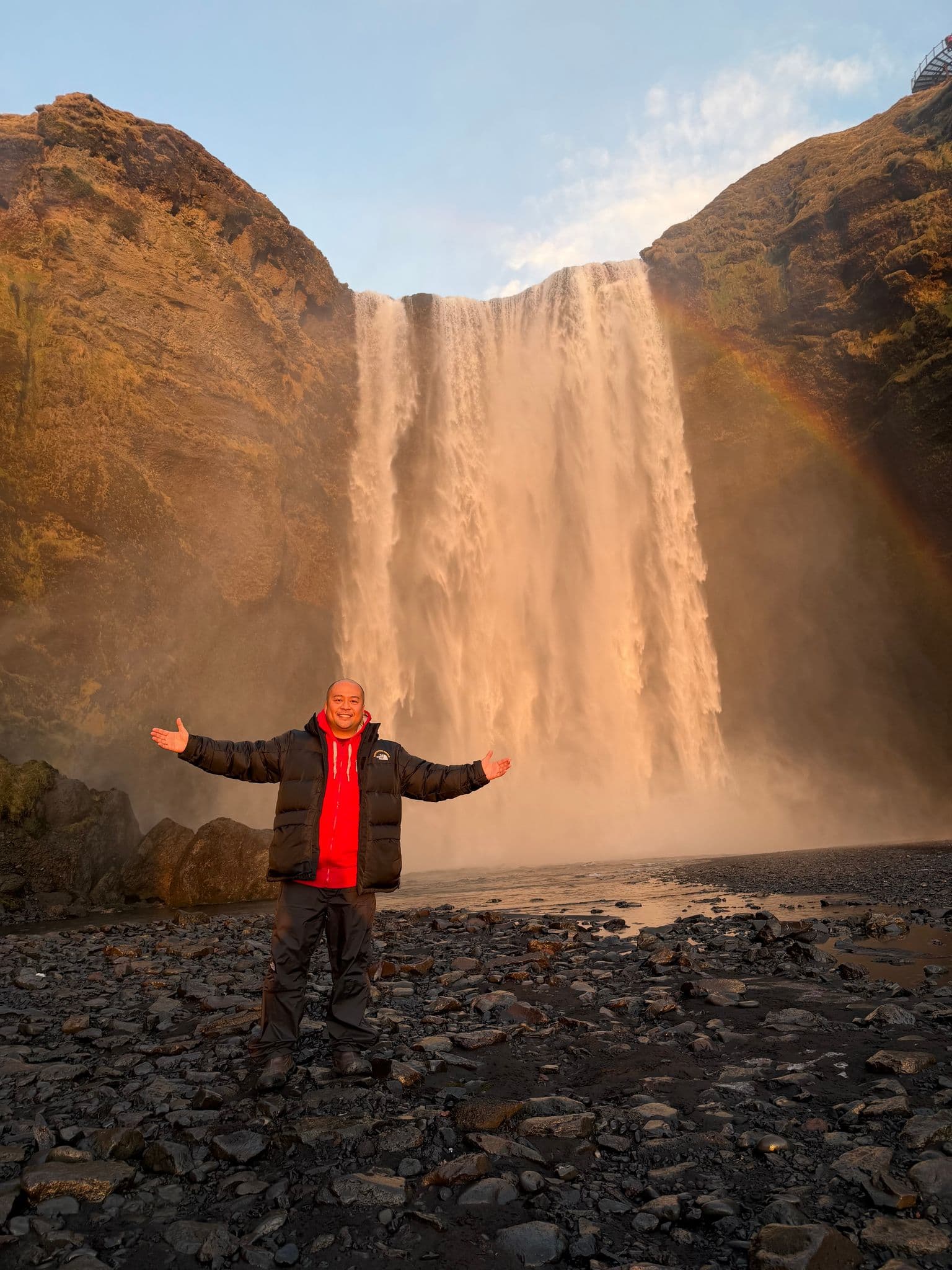 Skógafoss waterfall with a rainbow and a person standing on the rocky shore with arms outstretched, South Coast, Iceland