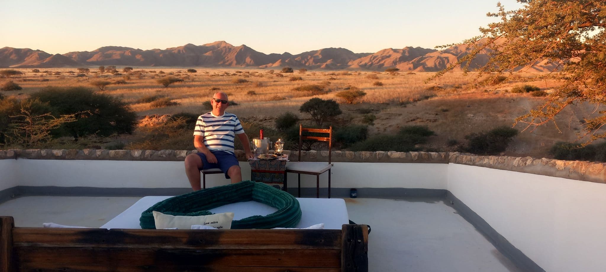 Rooftop sky bed and a man sitting with champagne overlooking the Namib Desert plains and distant mountains, Namibia.