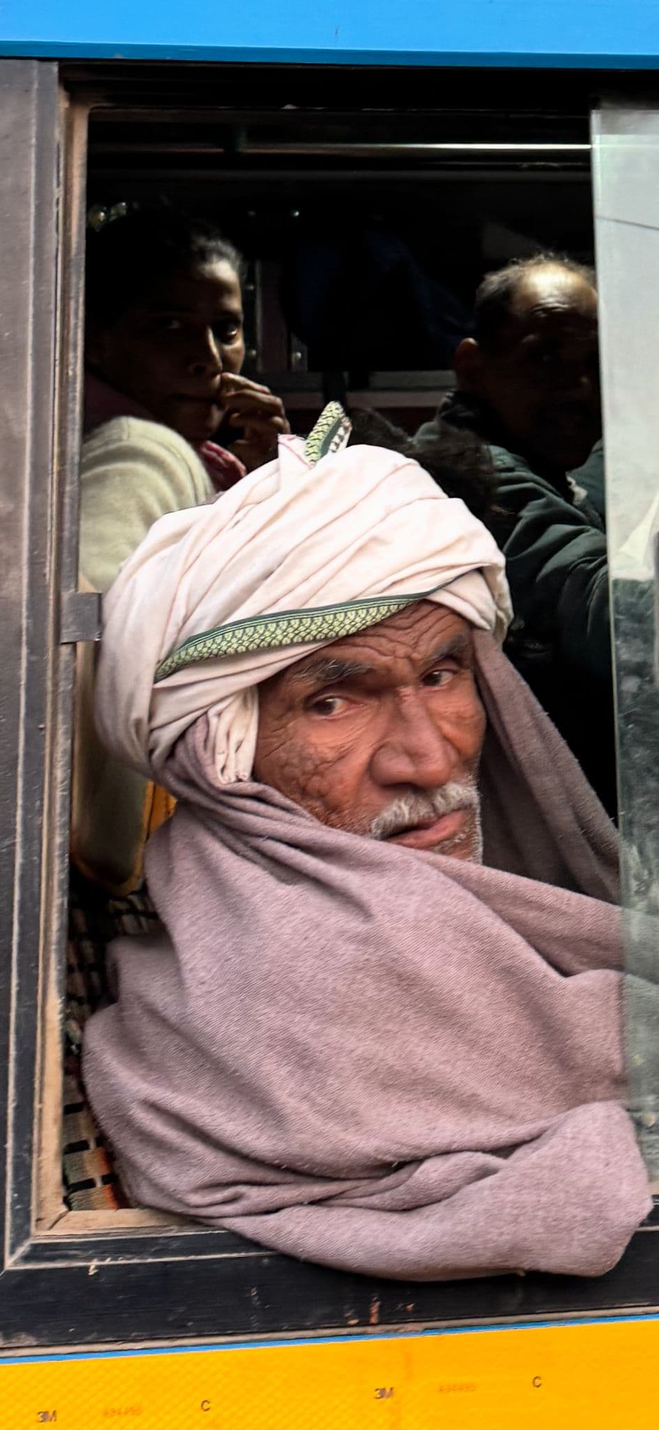 An older man in a turban wrapped in a shawl looking out a crowded bus window near Ranthambore National Park, India.