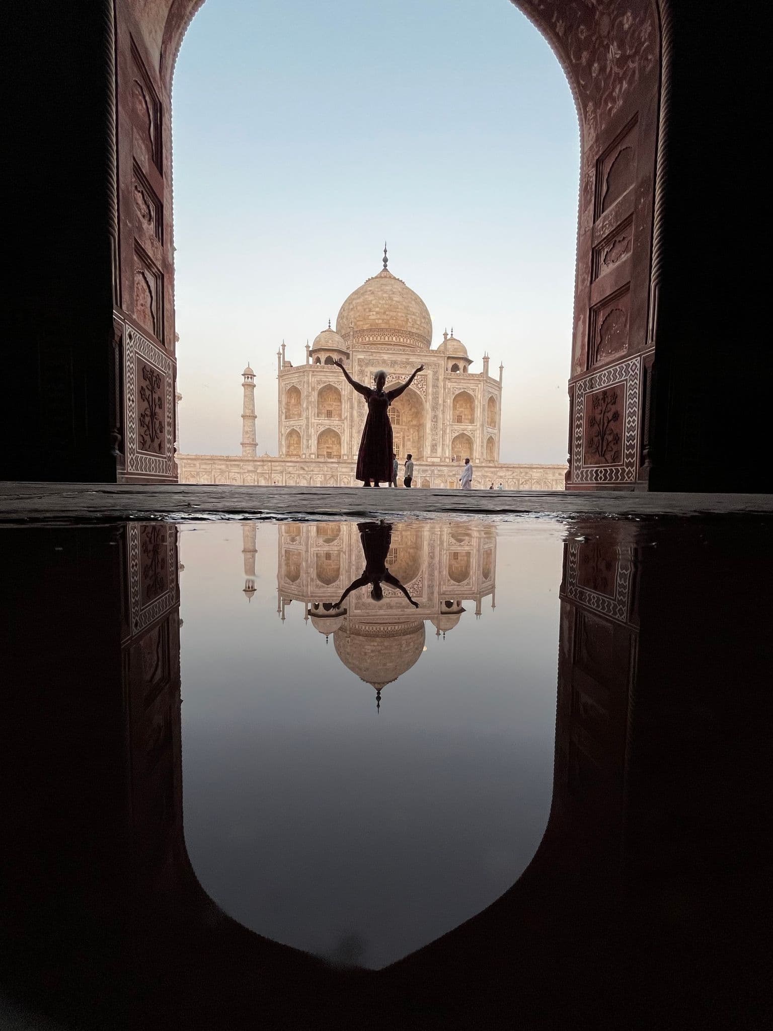 Taj Mahal framed by a carved arched gateway with a silhouetted person raising their arms and the monument mirrored in a foreground puddle, Agra, India.