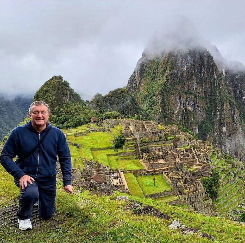 Machu Picchu ruins and terraces with Huayna Picchu behind, a traveler kneeling on a grassy overlook, Cusco, Peru