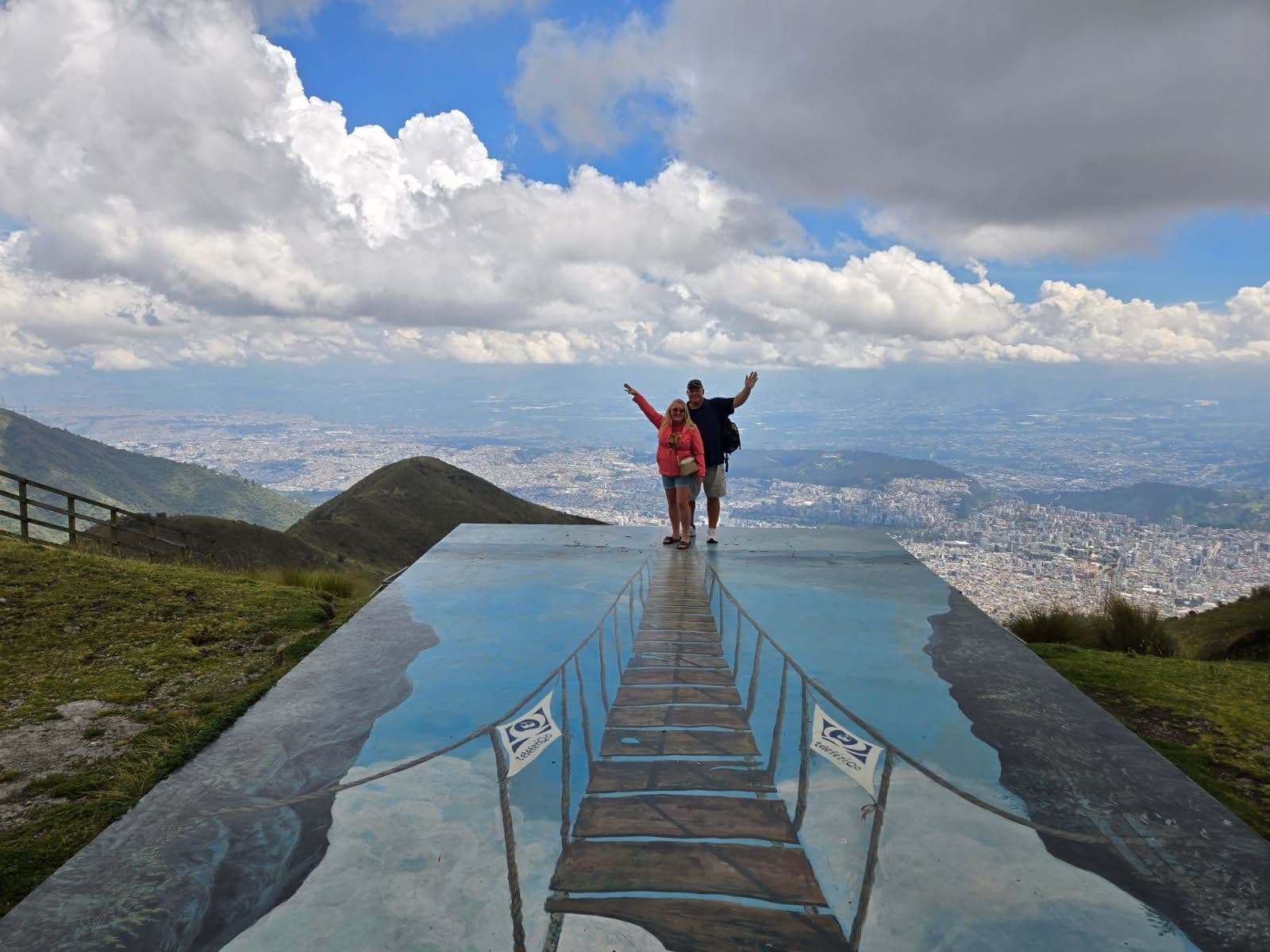 Painted platform showing a rope-bridge illusion leading to two travelers standing at the TelefériQo (Cruz Loma) viewpoint above Quito, Ecuador.
