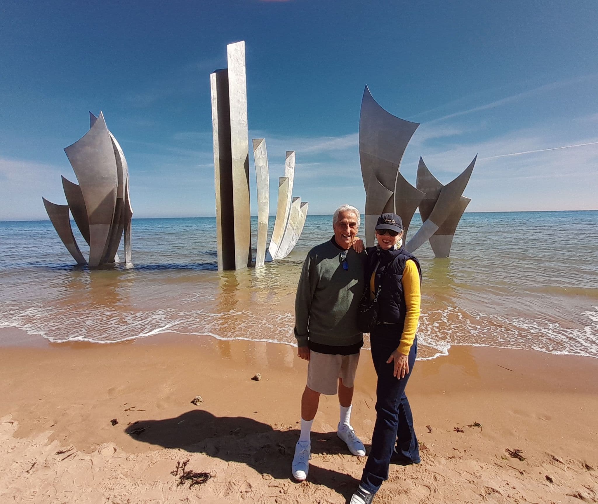 Les Braves sculpture at Omaha Beach with two travelers standing on the sand near the shoreline, Normandy, France.
