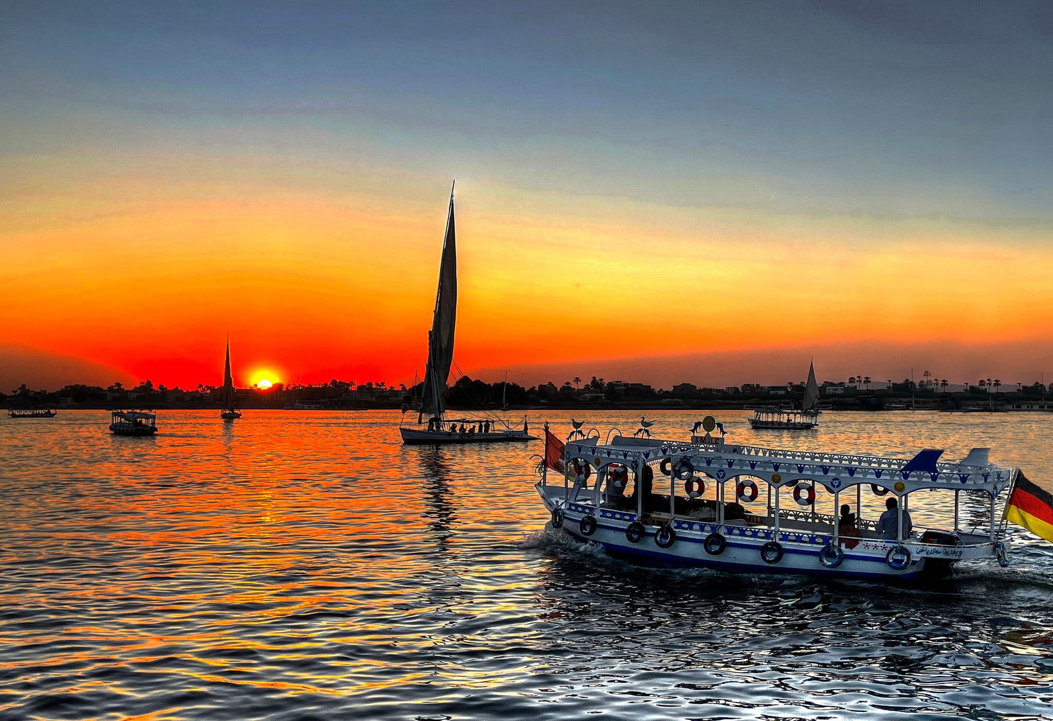 Nile River at sunset with a tall felucca and a decorated passenger boat sailing across the water, seen from a riverside hotel, Egypt.