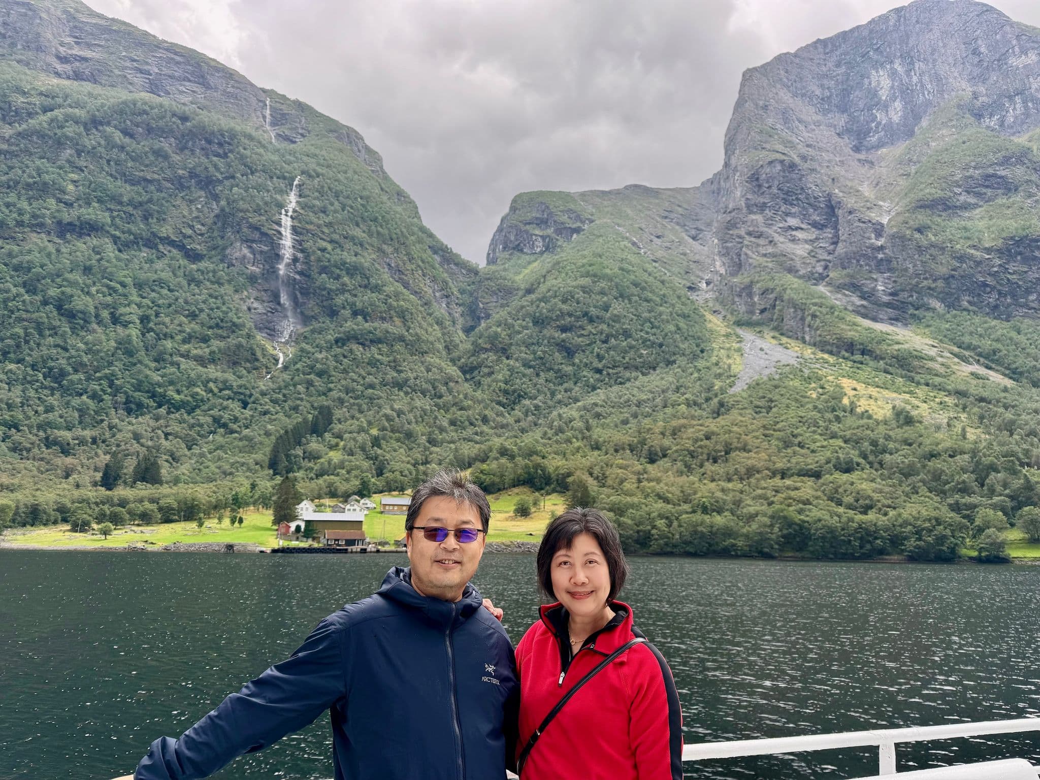 Nærøyfjord with a waterfall and steep green mountains, with two travelers standing on a cruise railing in Norway.